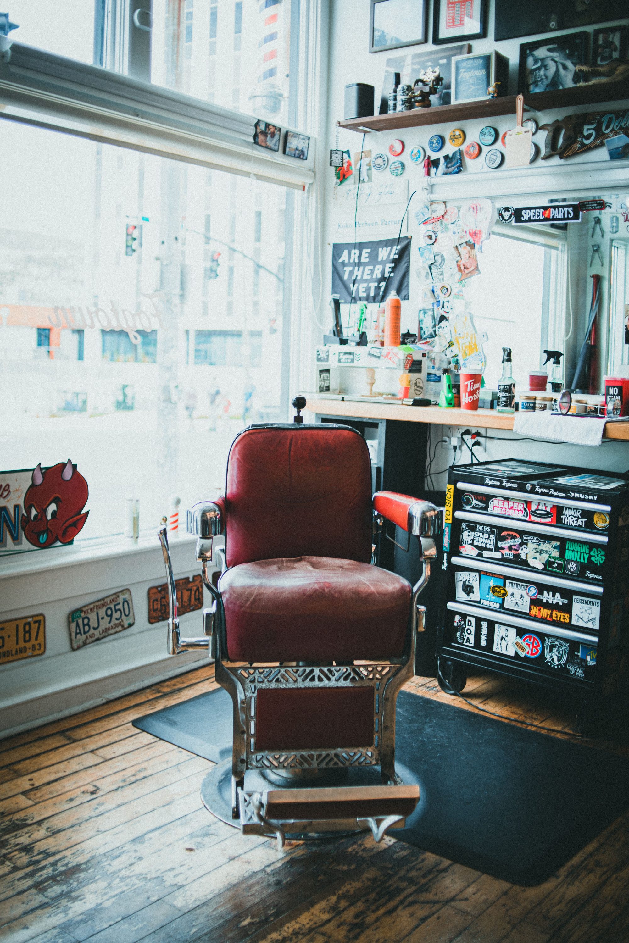 Vintage red leather barber chair beside a sunlit shopfront window, surrounded by decades of shop memorabilia and grooming products