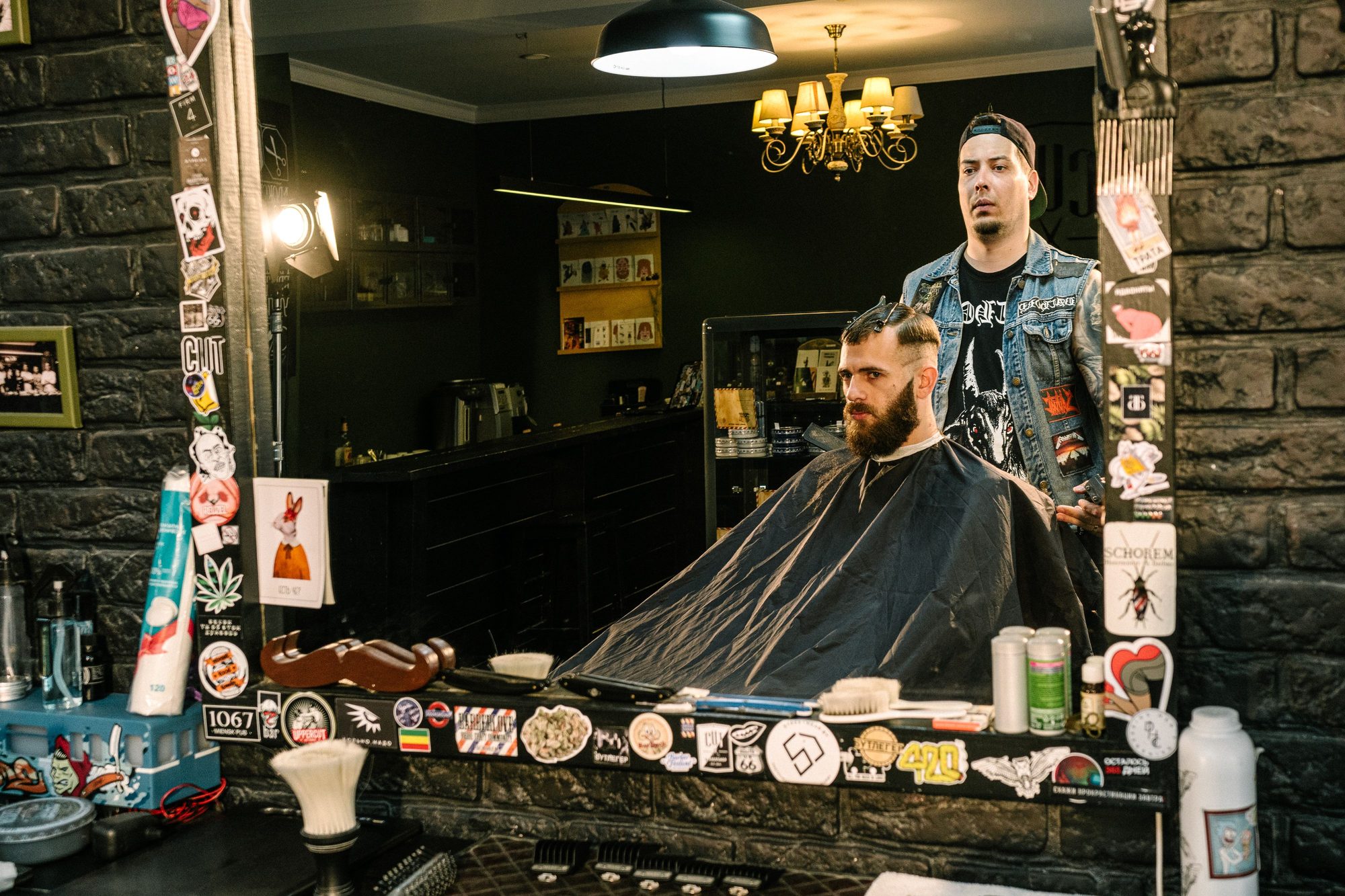 A barber giving a bearded client a fade haircut under warm chandelier light in a dim, rock-and-roll styled barbershop