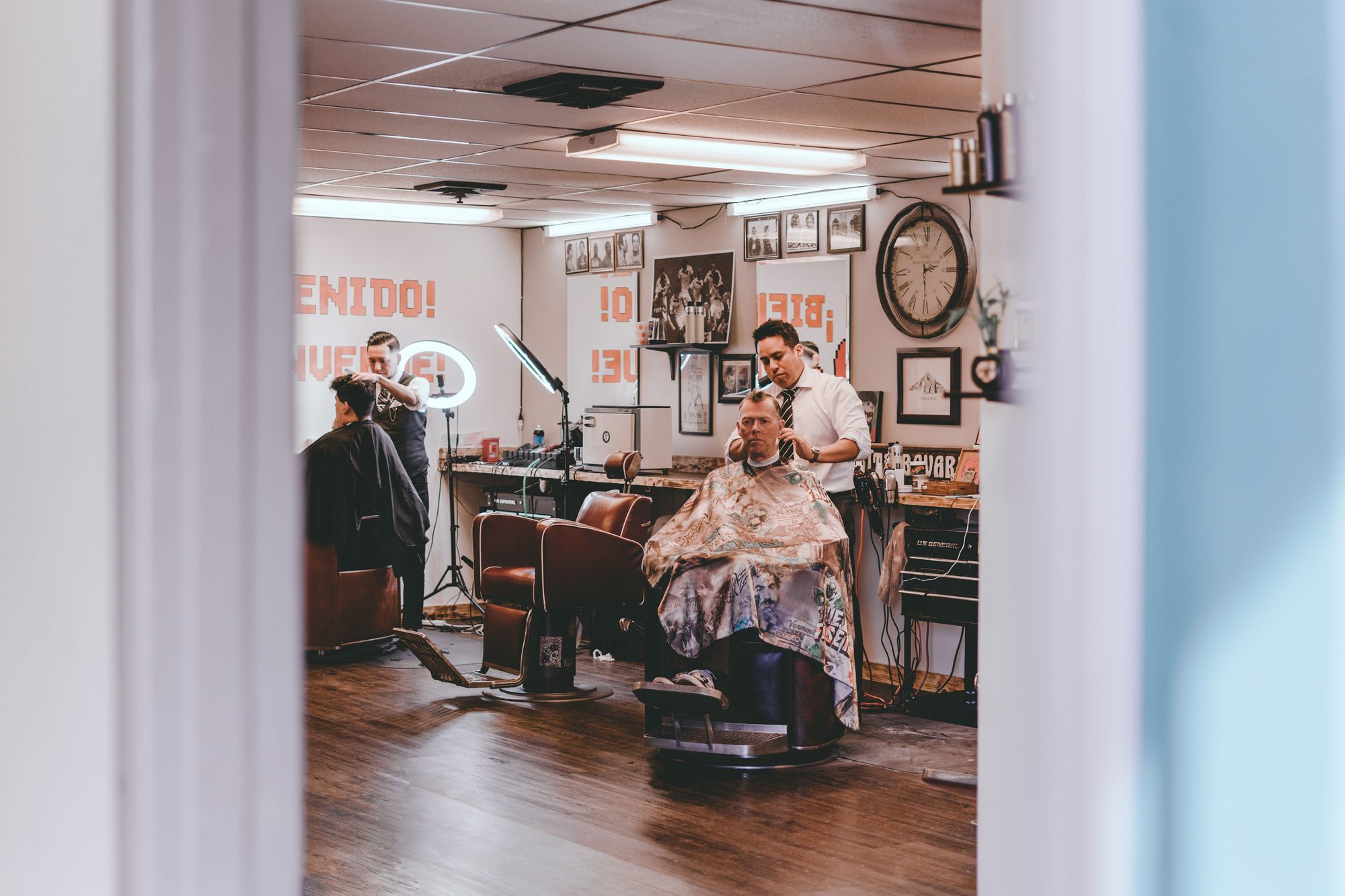 Two barbers working simultaneously on clients in a busy, light-filled barbershop with vintage chairs and a wall clock