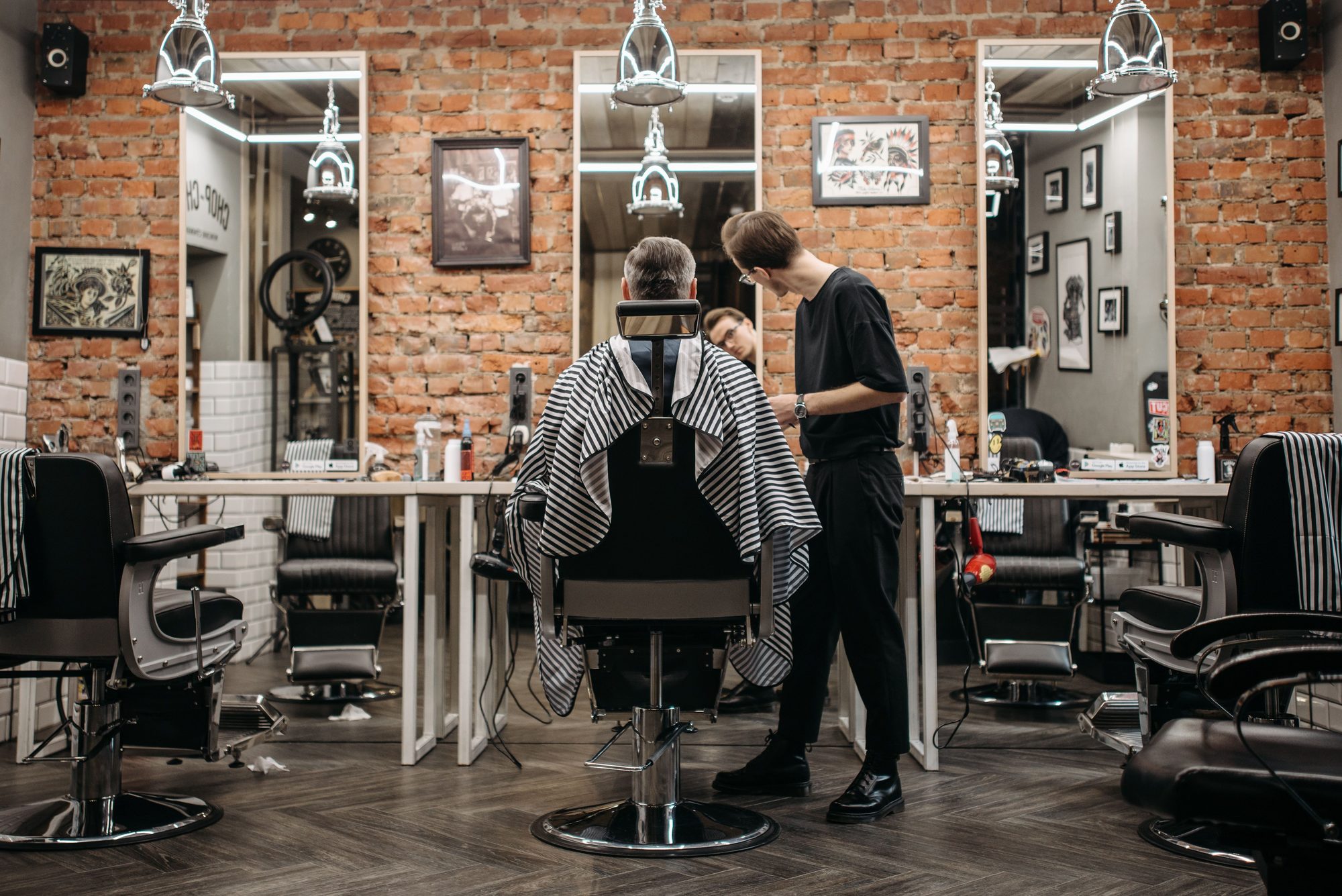 A barber trimming a client's hair at an exposed-brick grooming station flanked by mirrors and vintage pendant lamps